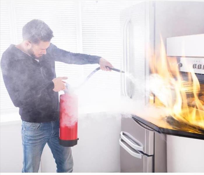 Young Man Using Red Fire Extinguisher To Stop Fire Coming From Oven In Kitchen